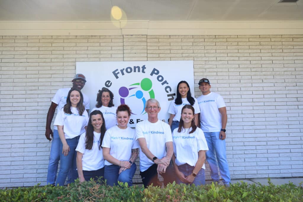 Group of smiling volunteers in white t-shirts posing in front of a banner on a brick wall, with greenery in the foreground.