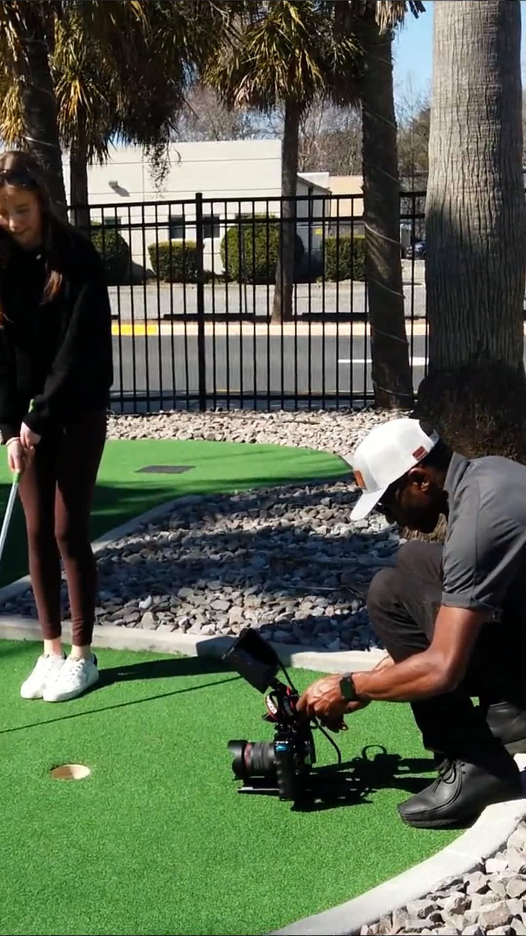 Cameraman kneels on a putting-green, filming a standing woman with a professional camera setup nearby.